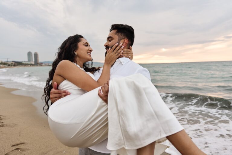 A man lifting his fiancée in the air while celebrating their engagement at the beach in Barcelona.