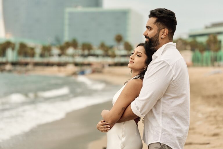 Groom-to-be hugging his fiancée from behind on a Barcelona beach with the city skyline in the distance.