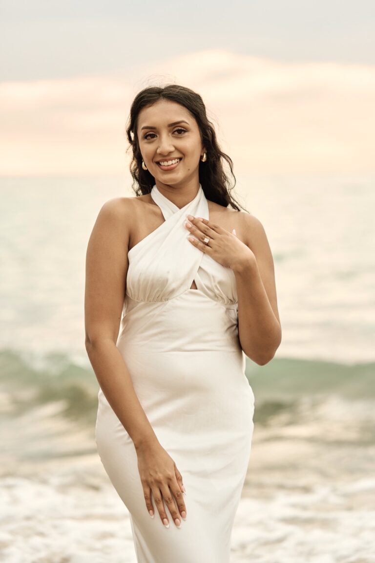 A happy woman smiling and showing her engagement ring during a sunrise session on a Barcelona beach.