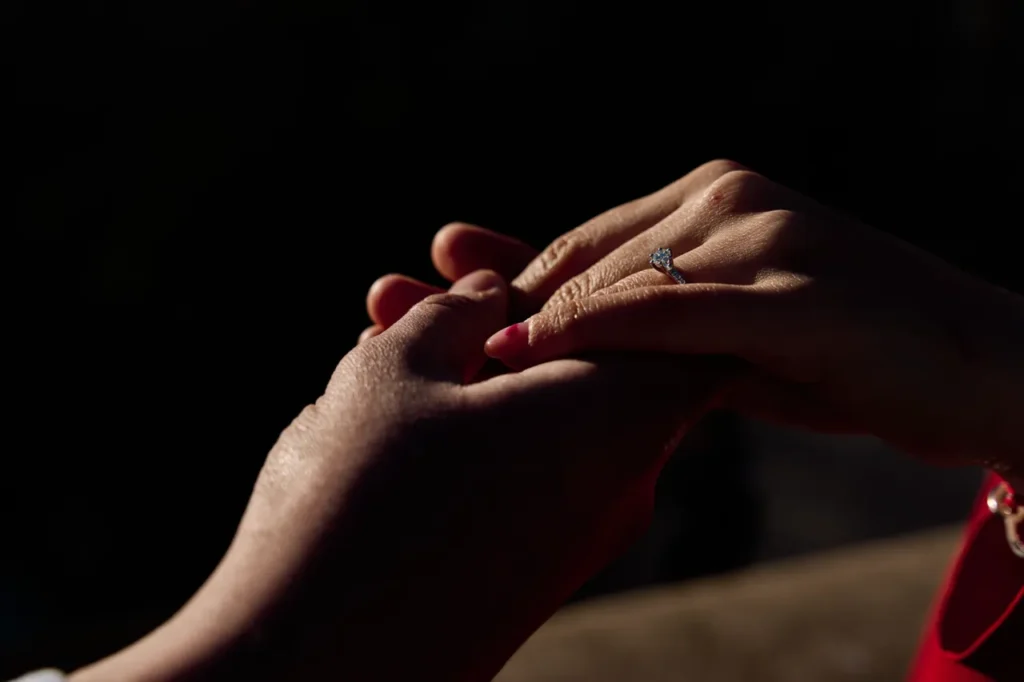 Artistic close-up of a man's hands placing a diamond ring on his fiancé's finger.