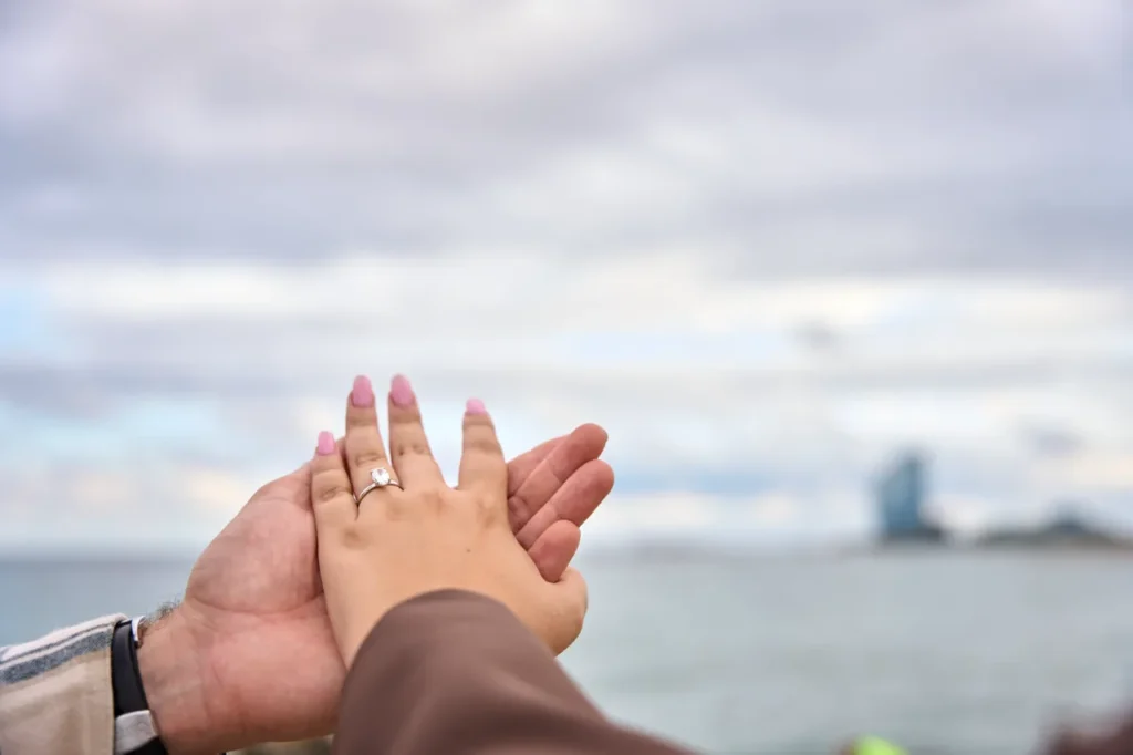 Close-up of a couple holding hands with an engagement ring by the Barcelona seashore.