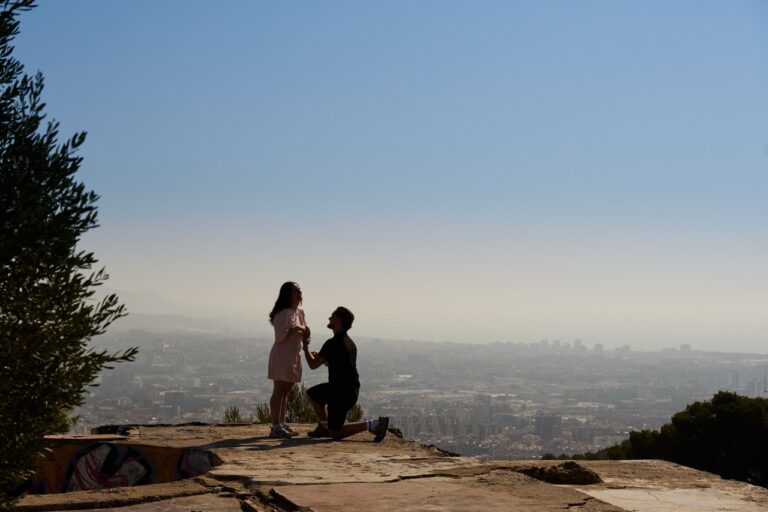 Young man proposing marriage to a surprised woman on one knee, silhouetted against the city skyline of barcelona
