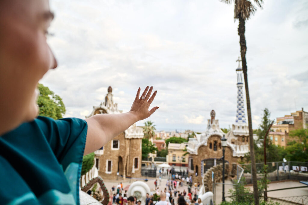Newly engaged woman raising her hand to show the ring, with the Park Güell pavilions behind