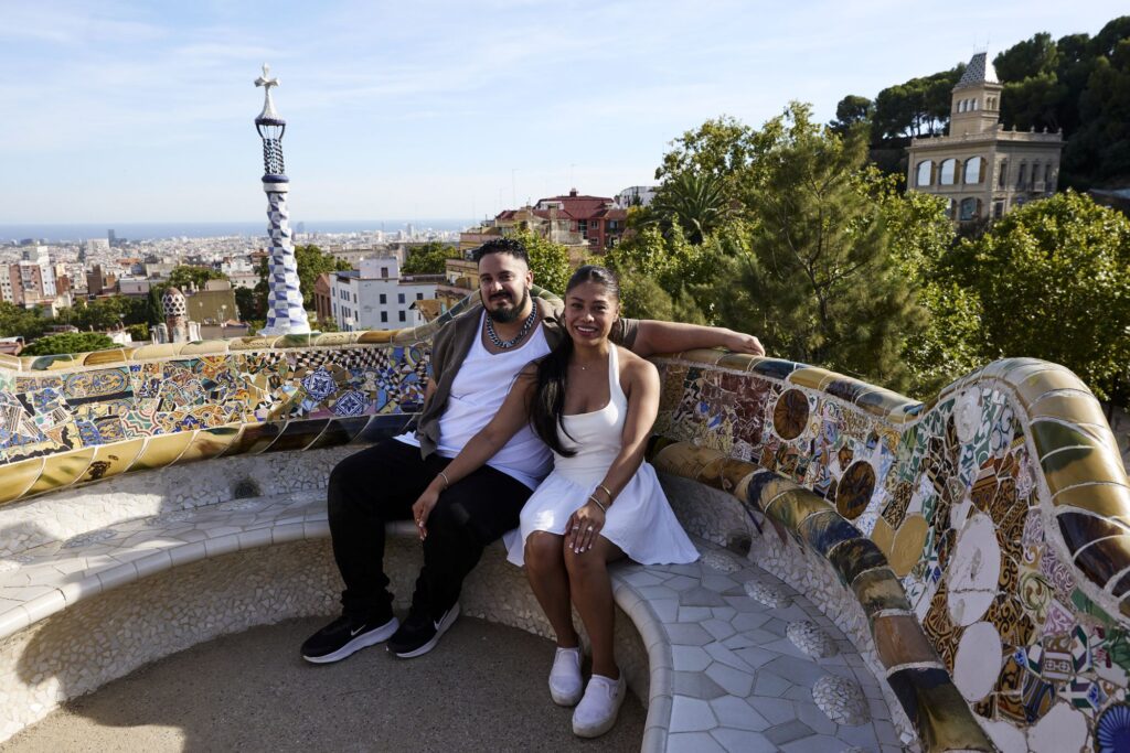 Couple sitting on the trencadís mosaic bench at Park Güell with Barcelona and the sea behind them